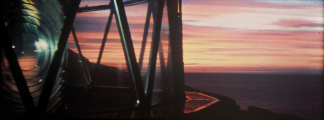 A still photograph taken from within the lamp section of a lighthouse at sunset; the globe of the unlit lamp is at far left, with a pink sunset in the distance.