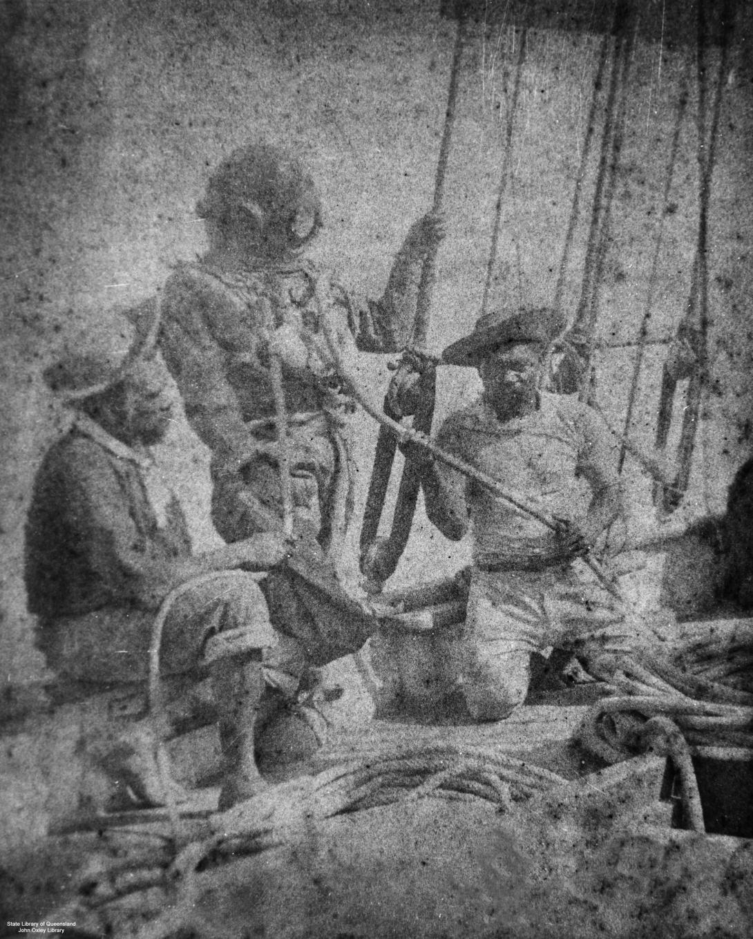 A black-and-white photograph from the late 1800s of pearl divers on a boat; one wears a diving helmet.