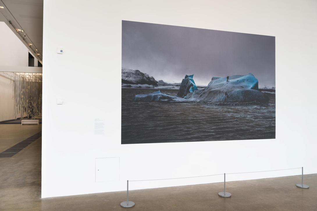 An installation view of a print on a white gallery wall; the work depicts a person standing on an iceberg, melting it with a blowtorch.