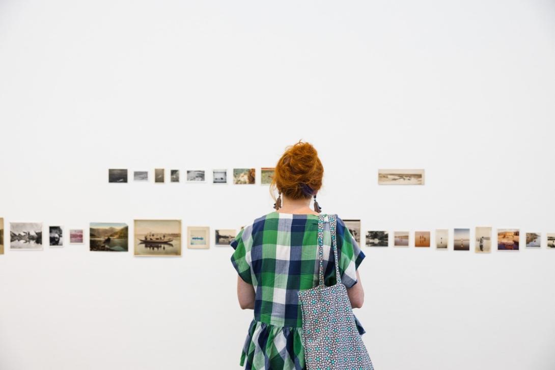 An installation view of many small works installed on a white gallery wall in two rows, with a feminine-appearing person in a blue-and-green dress looking on.