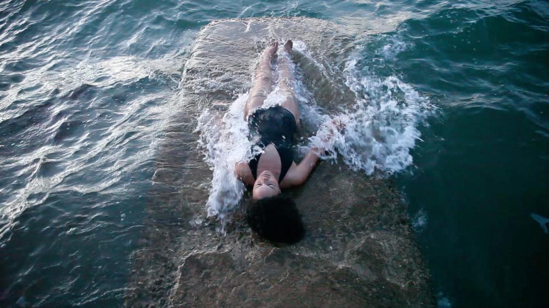 A photograph of a woman lying on a concrete outcrop in ocean water; she is wearing a black one-piece swimsuit and the water is washing up over her.