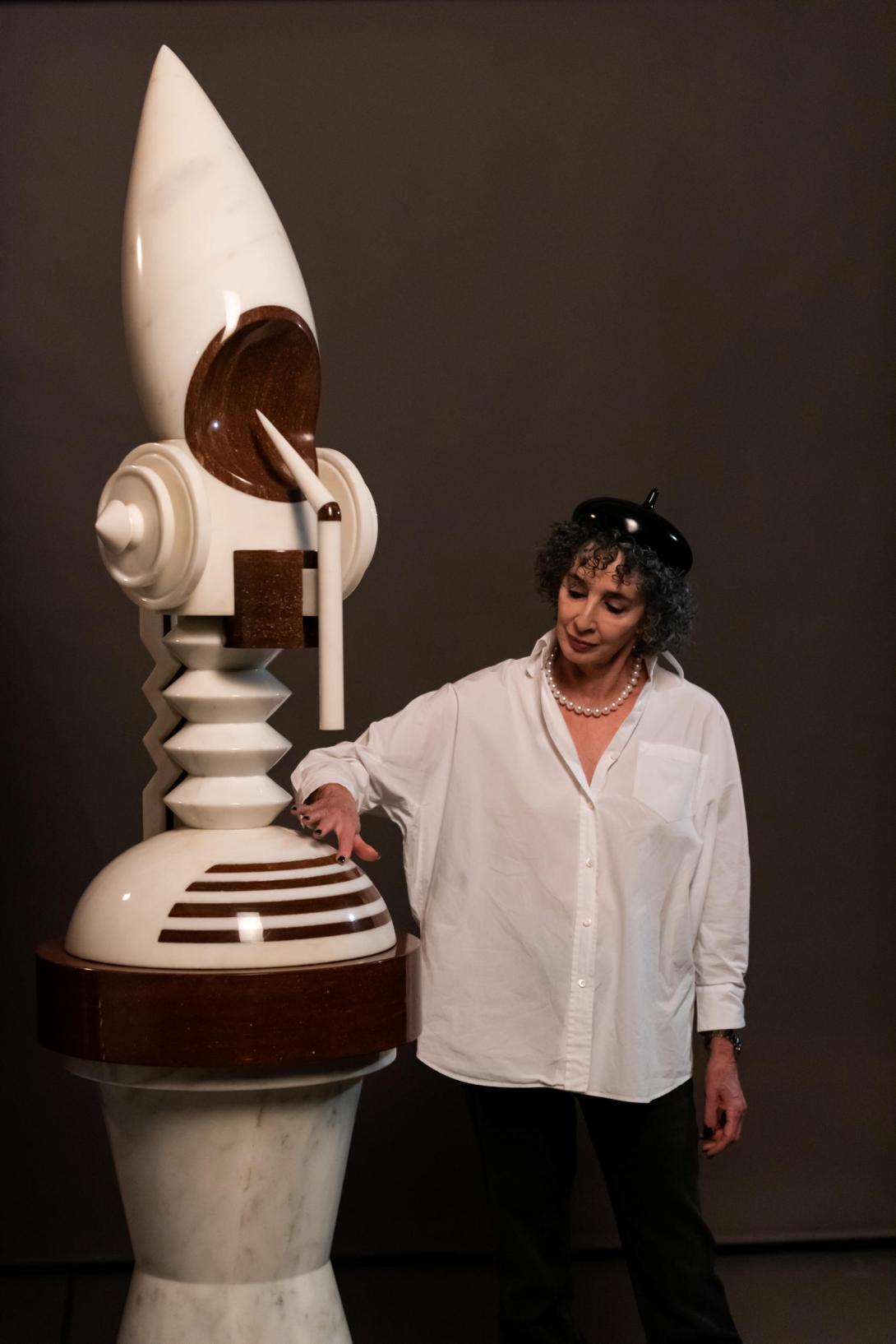 A woman wearing a white button-down shirt and a sculptural, shiny beret runs her hand over a larger-than-life marble statue, which takes the form of a modernist totem.