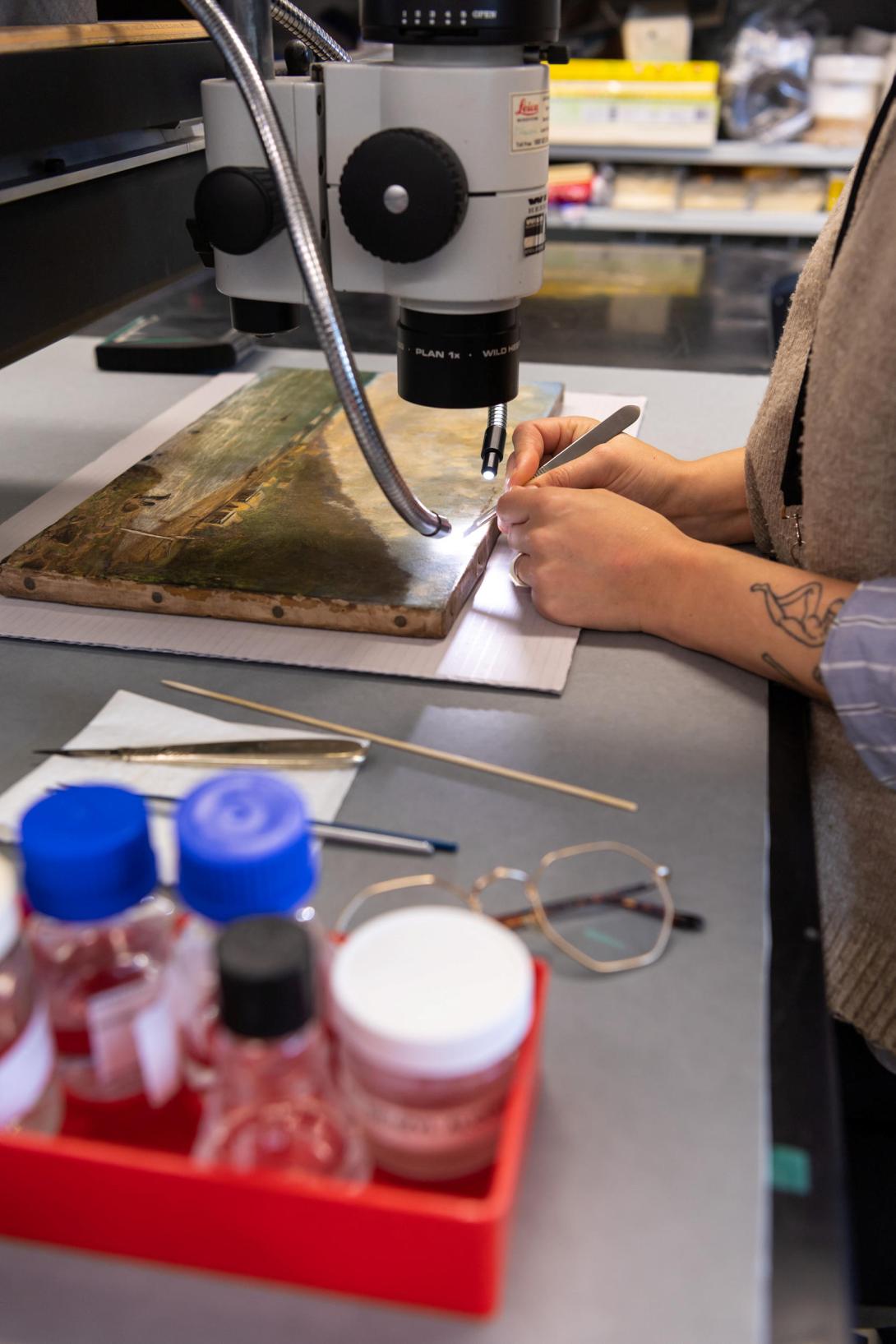 A conservator examines a painting under a microscope; we see only their hands, the equipment and, in the foreground, a red tray of jars containing various cleaning gels.