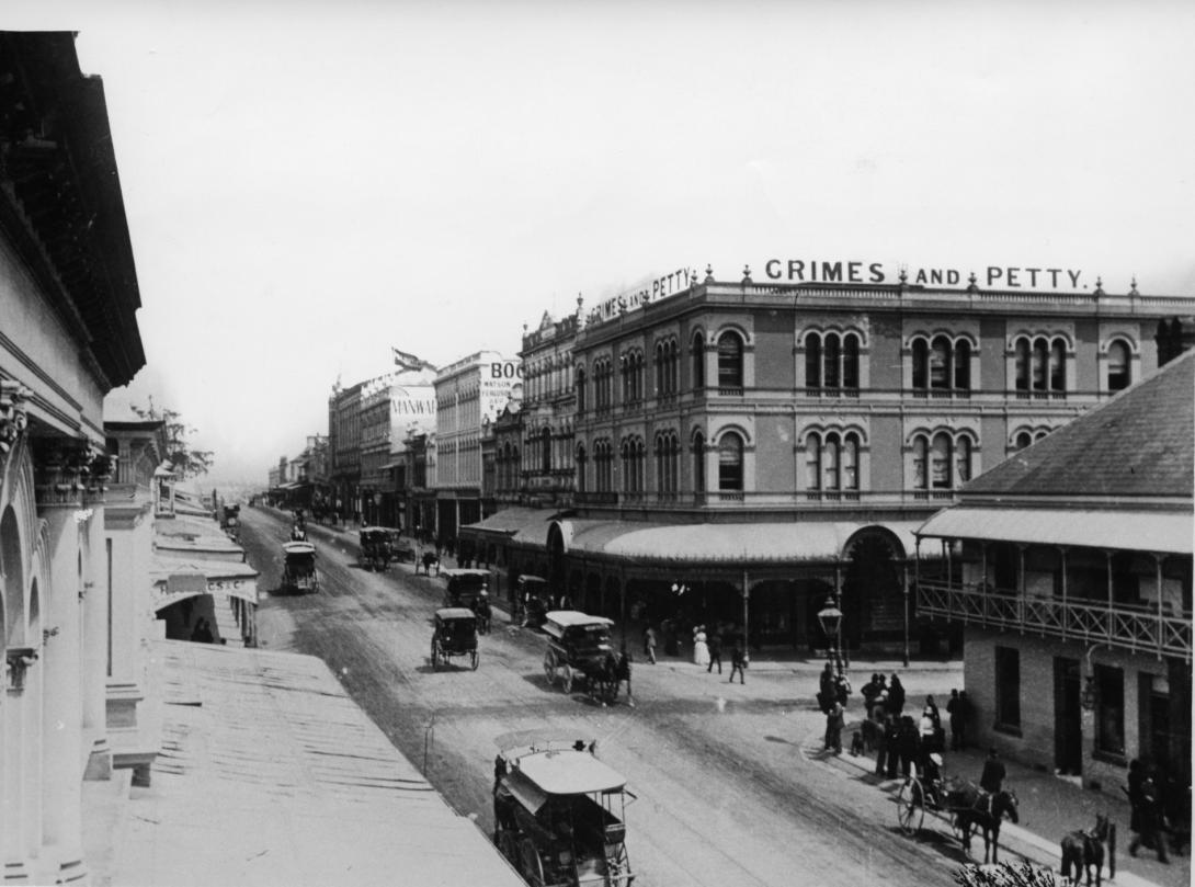A black-and-white photograph of a streetscape in which horse-drawn carriages can be seen passing shopfronts.