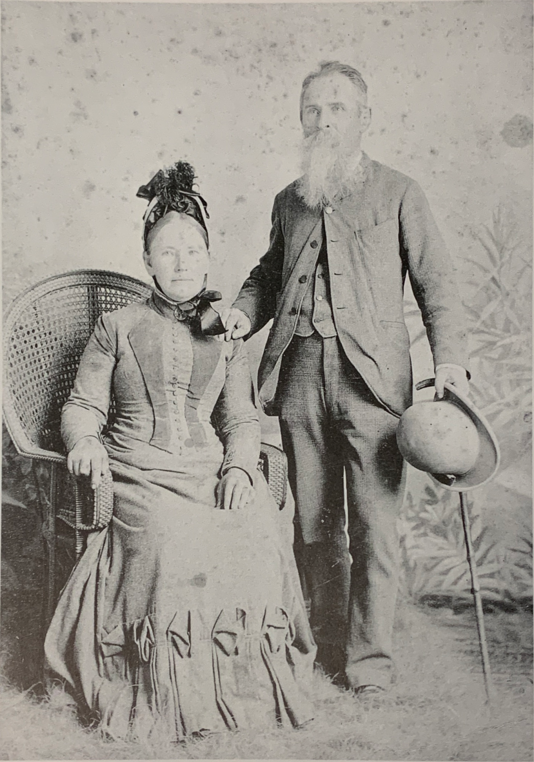 A black-and-white studio photo from the 1800s of a woman in period dress seated in a cane chair, with a man in a three-piece suit and carrying his bowler hat and cane standing beside her.