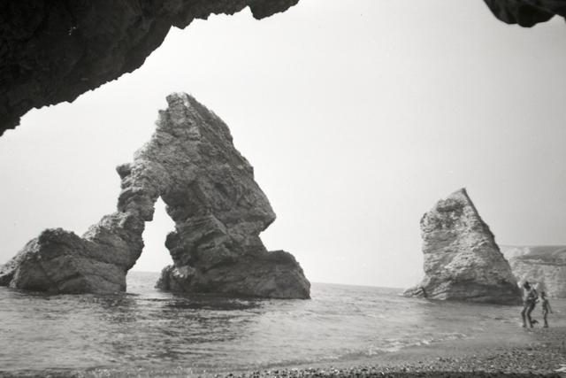 A black-and-white photograph of an arched rock formation in shallow ocean water, with two small figures walking past at bottom-right.