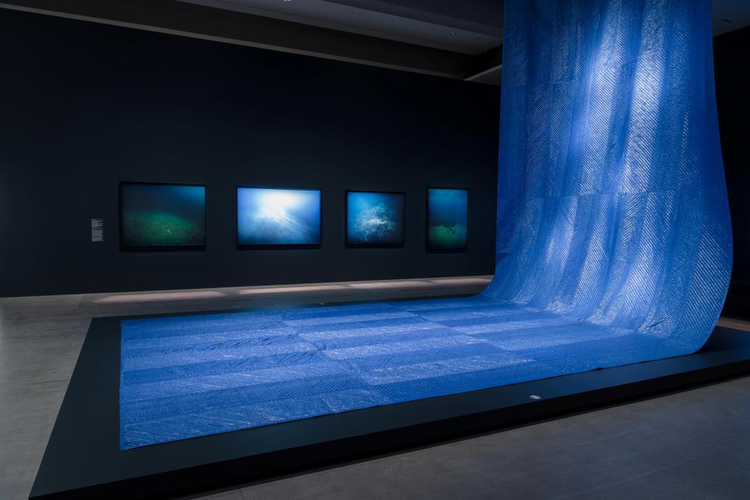 An installation view of a large, blue sculptural work that looks like water installed in a dark gallery space.