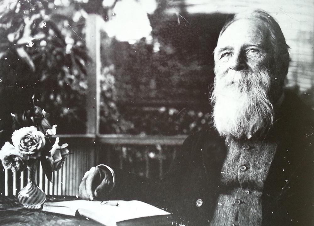 A black-and-white photograph of an older man seated on a verandah in the late 1890s; he is smiling gently, and seated with his hand at an open book and with a small vase of roses at the table.