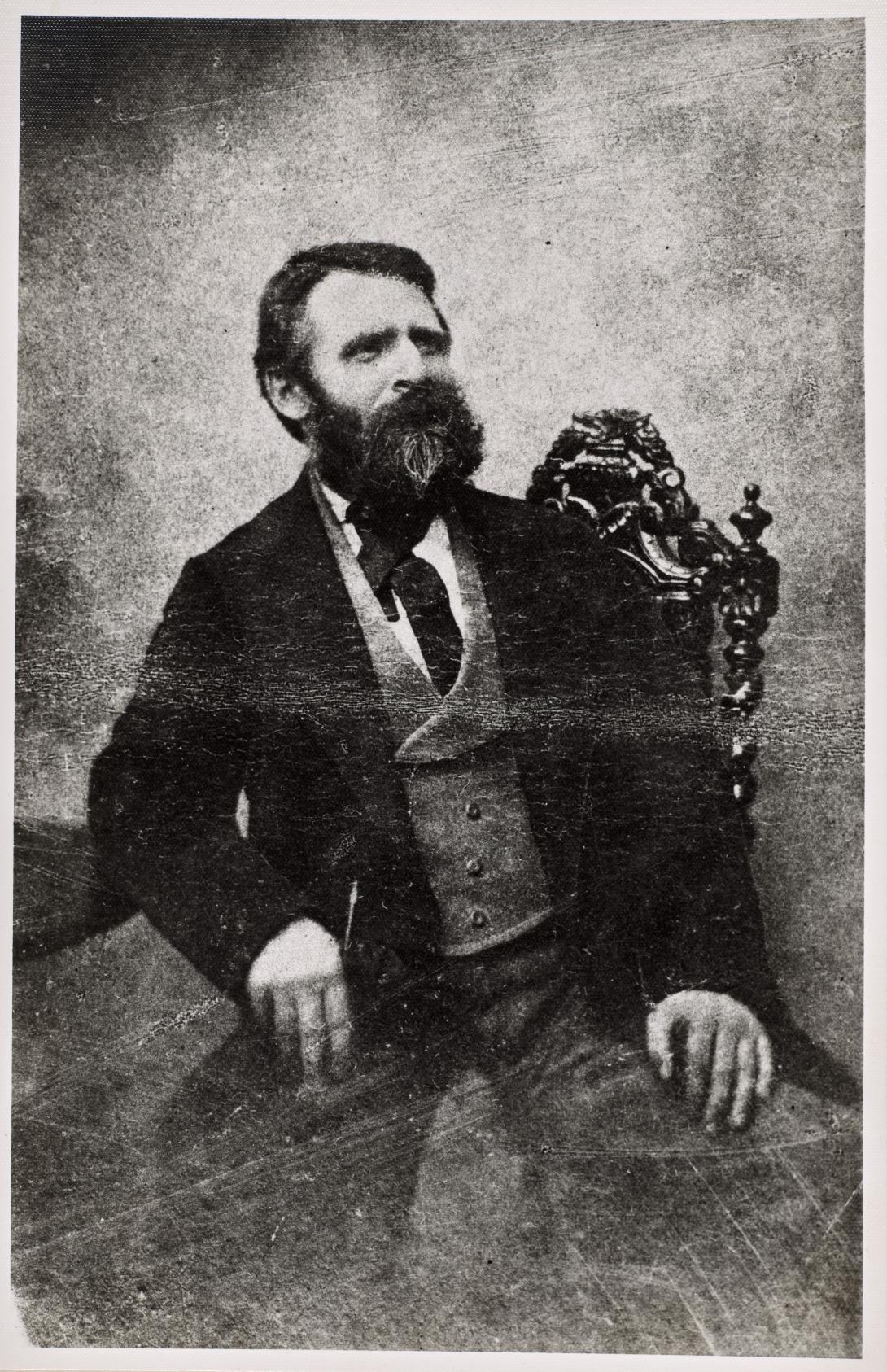 A black-and-white photograph of a man wearing a waistcoat, tie and coat, seated on an ornate chair.