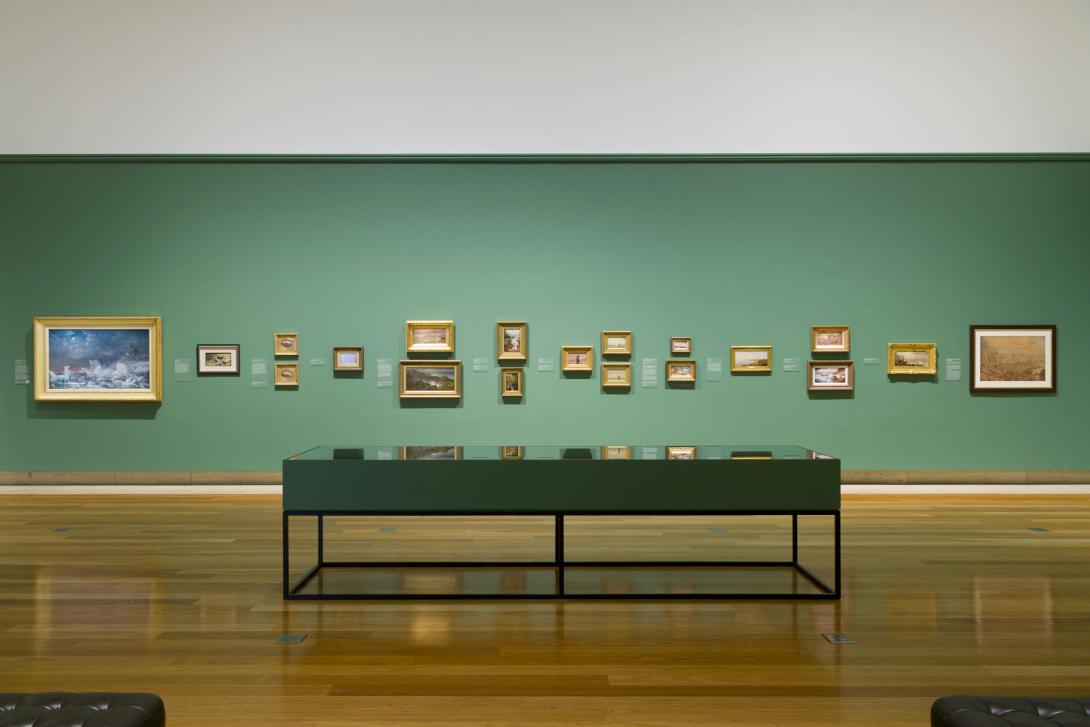 An installation view of small works of art hung on a green gallery wall, with a glass cabinet in the foreground.
