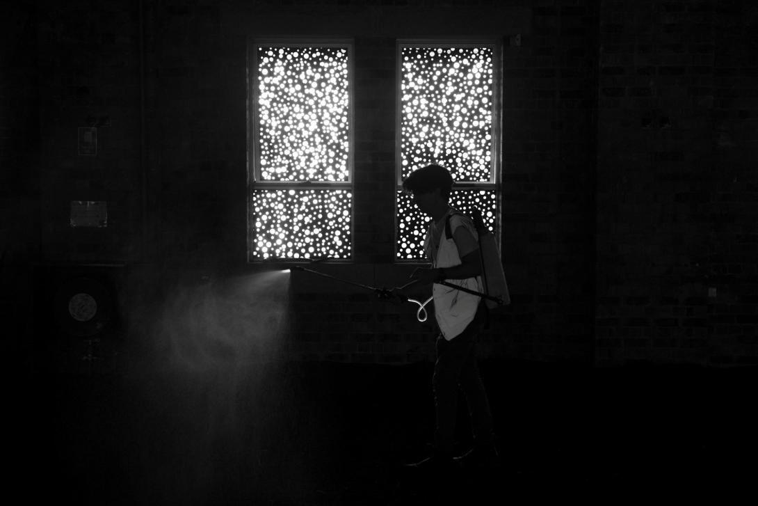 A very dark installation view in which light filters softly through a window treatment; a figure sits in the window and we see their silhouette.
