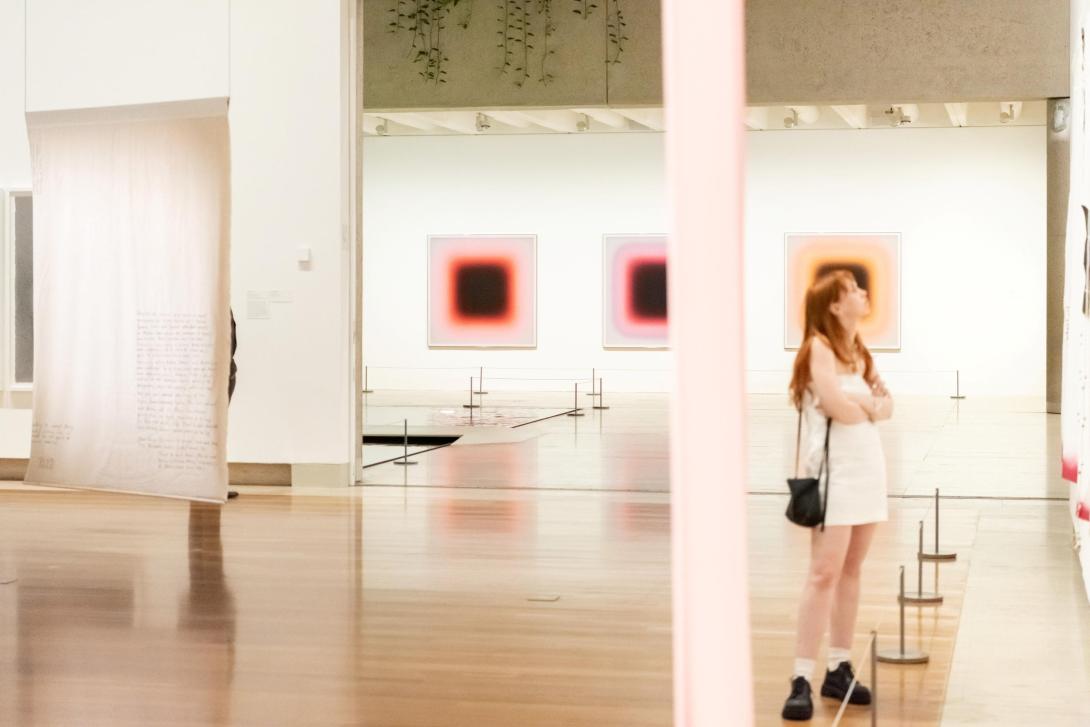 An installation view of a bright gallery space, with a suspended work seen from the verso side; works hung on a wall in the far background; an a visitor looking at a work out of our sight at the right-hand side.