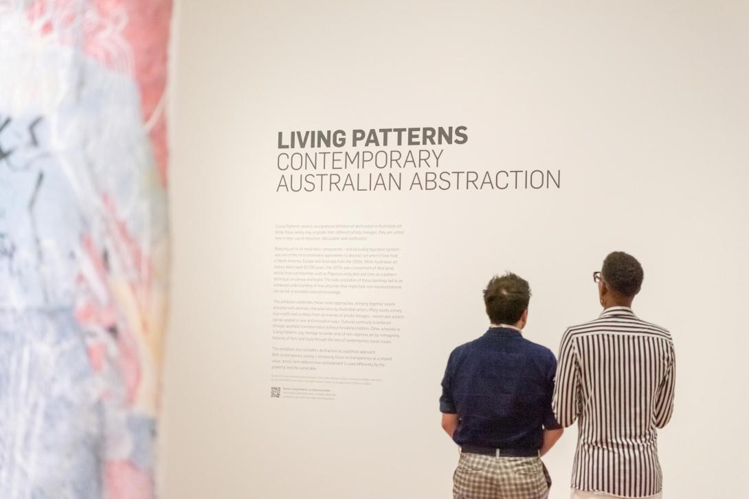 An installation view featuring a detail view of an artwork at left, a didactic on a white wall, and two visitors looking on.
