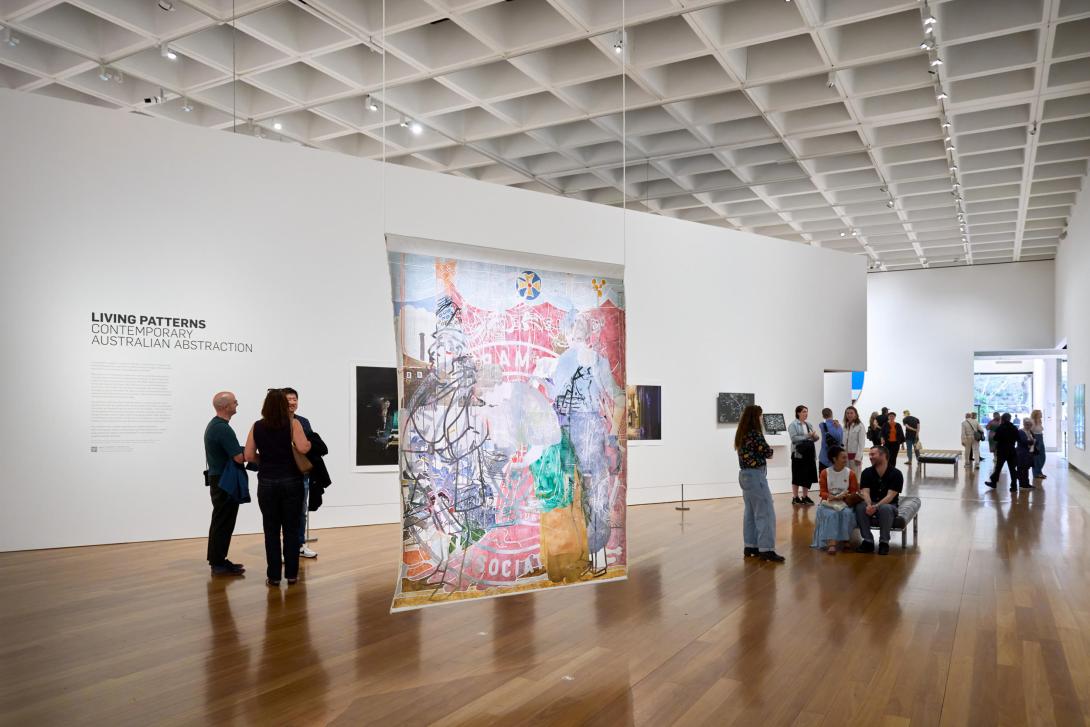 An installation view of a gallery space with many visitors; a colourful fabric work is suspended in the foreground.