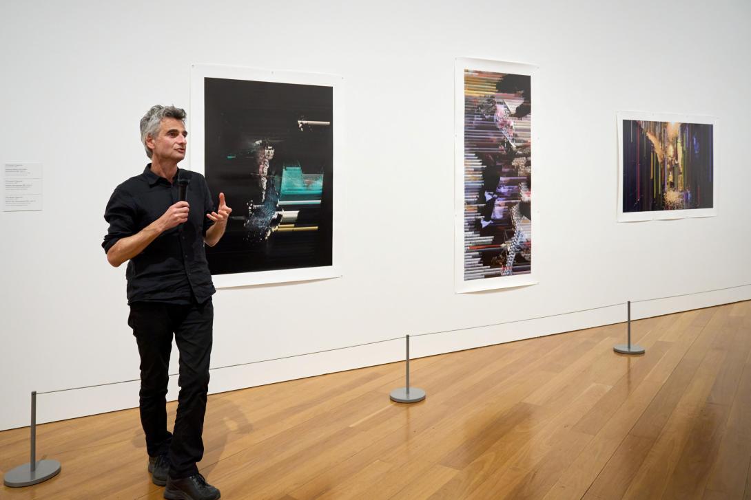 An artist dressed in black and with short, dark grey hair introduces the three prints installed on the white gallery wall behind him.