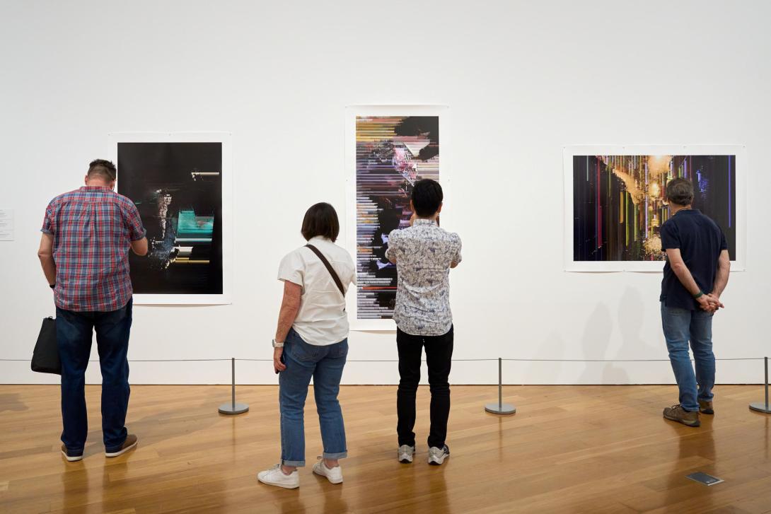 An installation view of three prints on a white gallery wall, with four people standing closely, looking on.