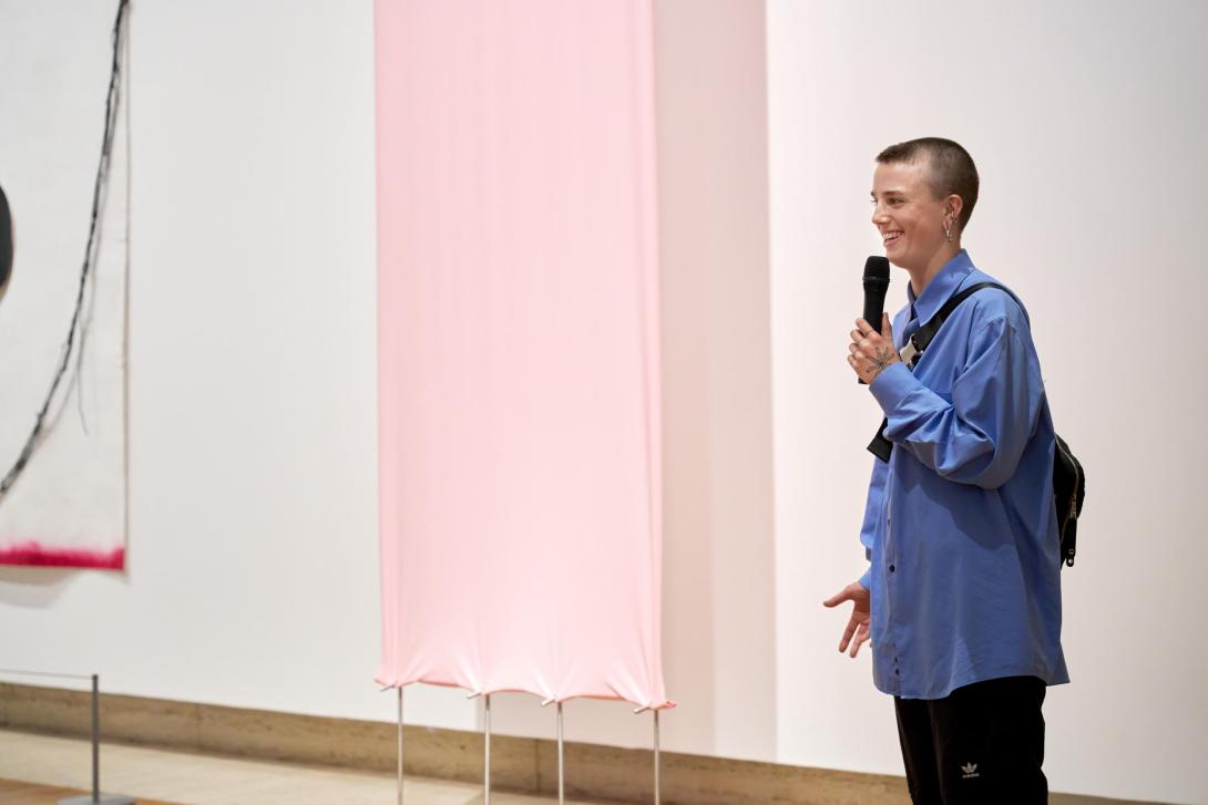 An artist with cropped hair and wearing a blue shirt stands to the right of their artwork, which is made of a sheet of pink latex held under tension.