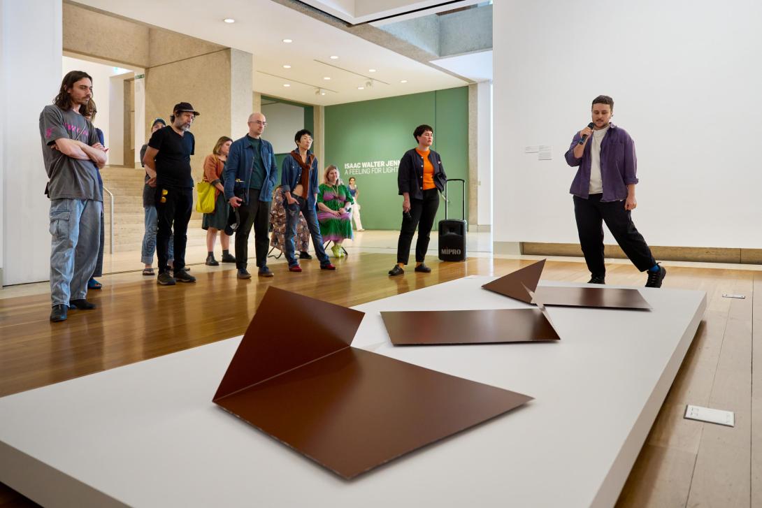 A photograph of three steel abstract sculptures installed on a low platform; standing behind these, the artist introduces their work to a tour audience.
