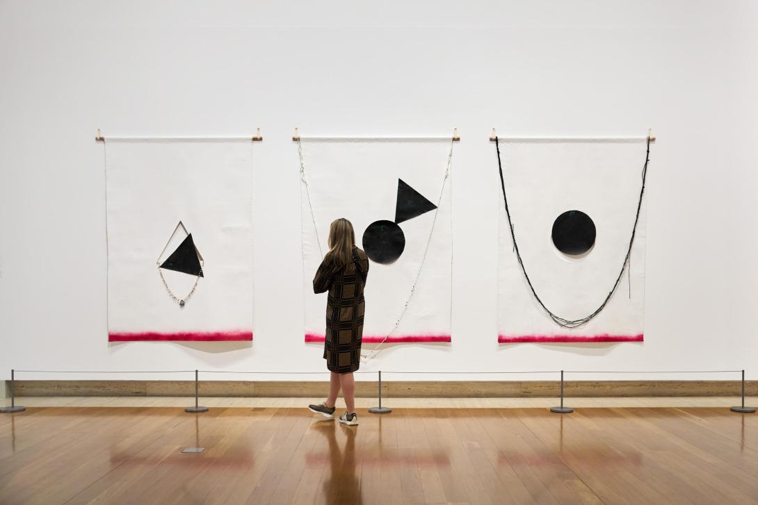 An installation view of three large, fabric works installed on a white gallery wall, with one visitor looking on.