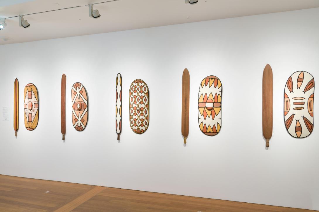 An installation view of five sets of shields and swords displayed on a white wall in a gallery space.