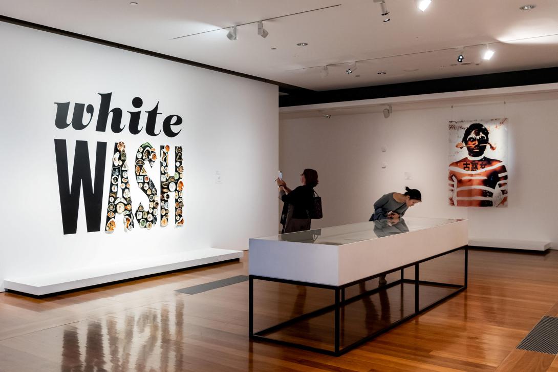 An installation view of a gallery space; on the left, a work reads 'whiteWASH', while an artwork depicting an Indigenous Australian man on the far wall. Two visitors look on.