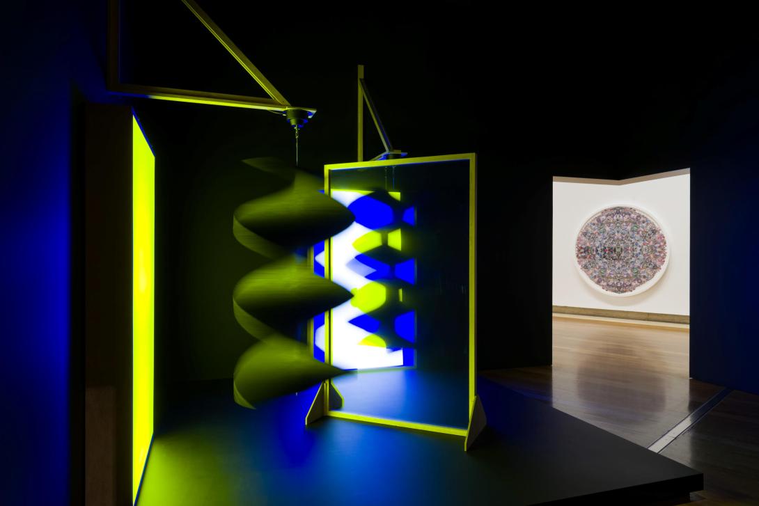 An installation view of a kinetic sculpture glowing yellow and blue in a dark room, with a view of another work on the wall in the background.