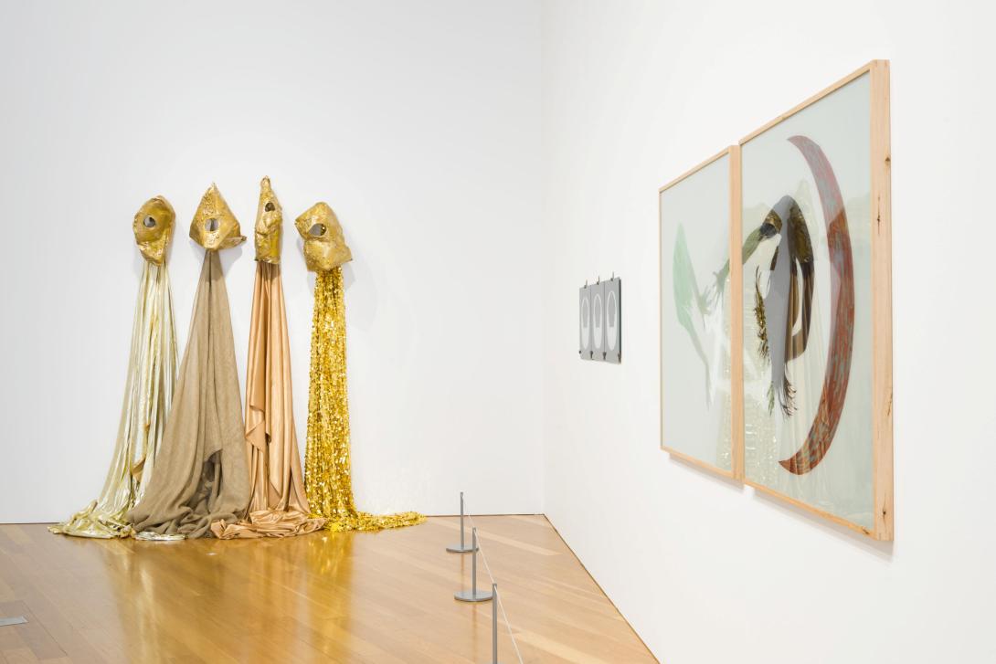 An installation view of a white gallery space with, at left, a gold-toned fabric work installed on the wall, with a diptych hung on the wall at right. 