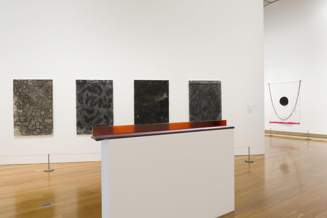 An installation view of a gallery with several abstract artworks installed: in the foreground, a sculptural work; behind that, four dark paintings hung on a white wall; and, in the distance, a further work hung on a wall.