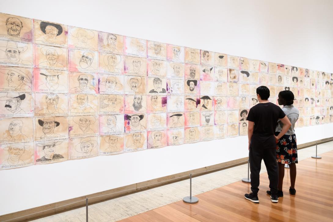 An installation view of a large work composed of many smaller drawings and collages on a white gallery wall, with two visitors looking on.