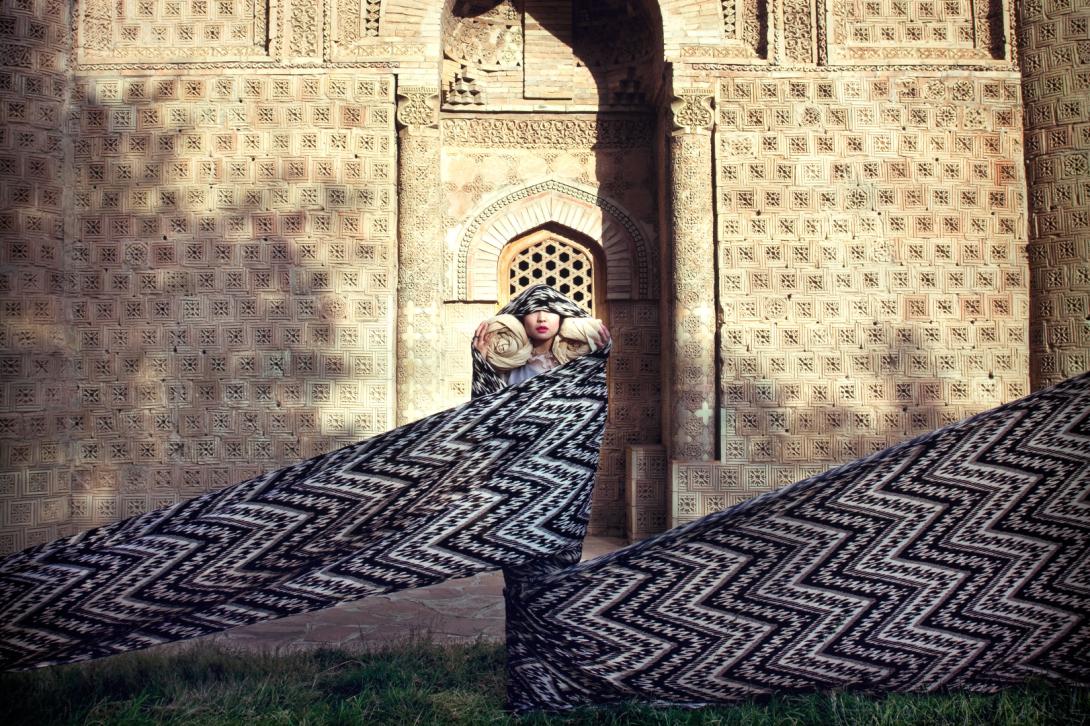 A still photo depicting a person standing before a stone palace-type building, wrapped in a long bolt of patterned fabric that spans the full length of the photo.