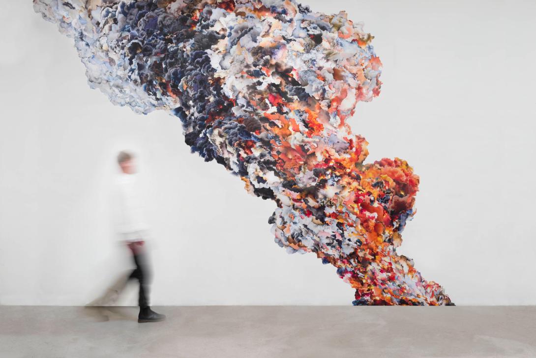 An installation view of a colourful work composed of many plumes of smoke collaged together, with a gallery visitor looking on at left.