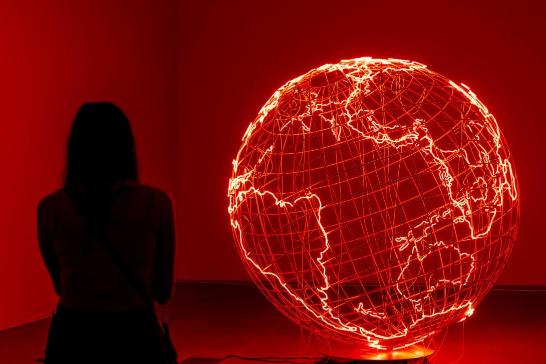 A glowing sculpture in the shape of a world globe installed in a gallery space, glowing red.