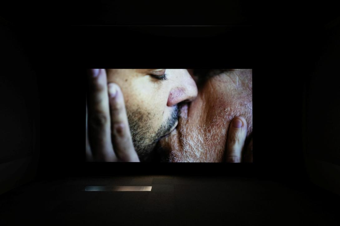 A still installation view of a video screening; two men kiss in the video, their hands tenderly touching one another's faces.