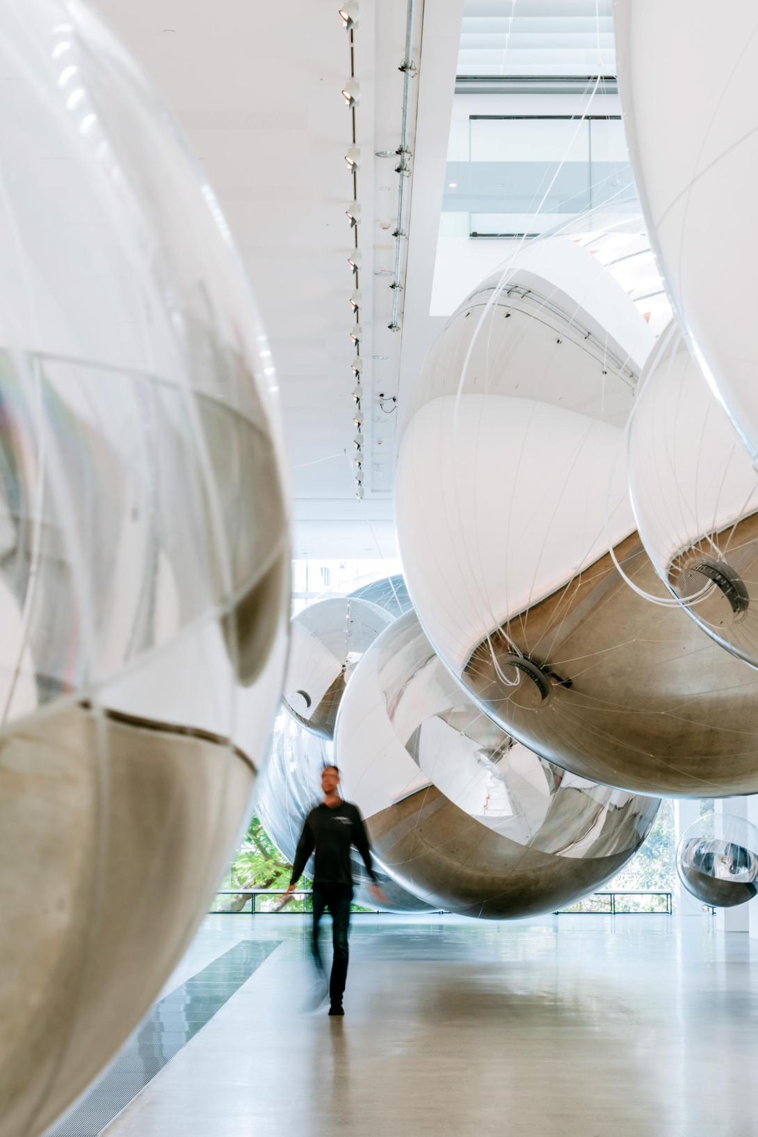 An installation view of enormous silver spheres suspended in a gallery space, with visitors looking on.