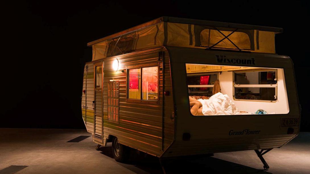 A view of a caravan installed in a dark gallery space; it appears to be occupied by two sleeping figures.