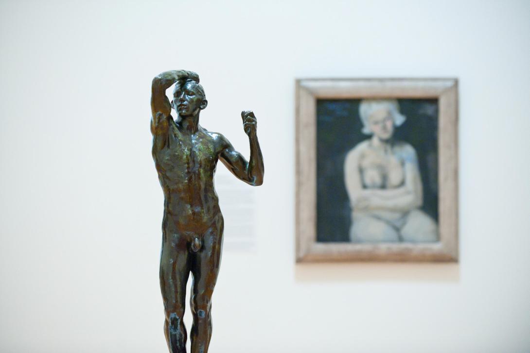 An installation view featuring a bronze statue of a man in the foreground, and an oil painting of a nude woman wearing a bonnet in the background.