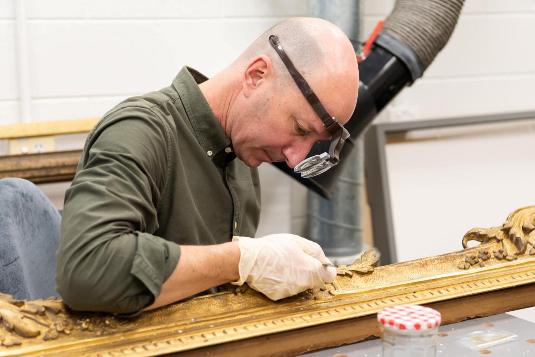 A man in a green long-sleeved shirt, magnifying loupes and latex gloves performs restoration work on a gilded frame, with an exhaust fan behind him.