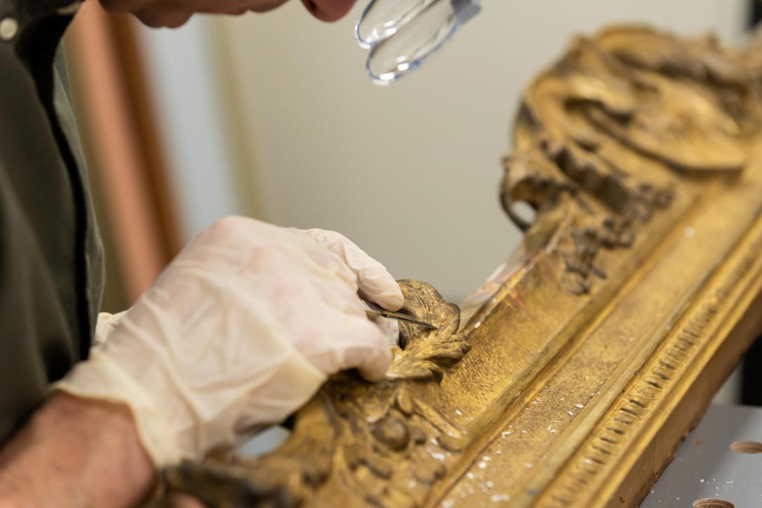 A gloved hand belonging to a frame conservator works on a gold-gilded frame.