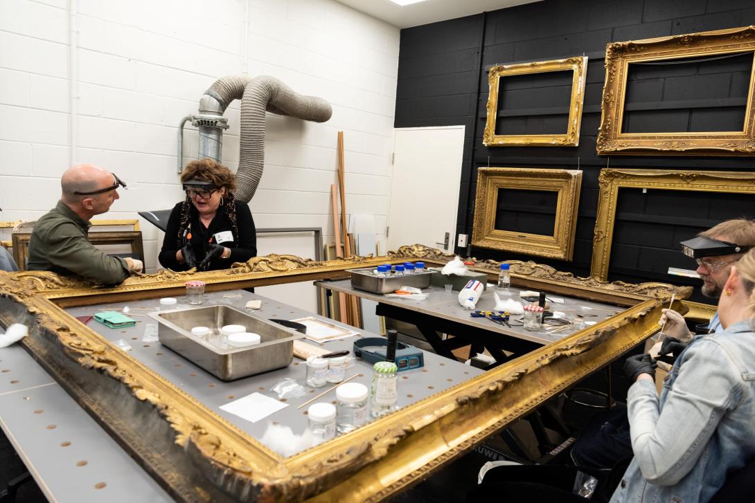 A photograph of four people seated around a large gilded frame, laid flat for restoration in the QAG Framing Workshop.