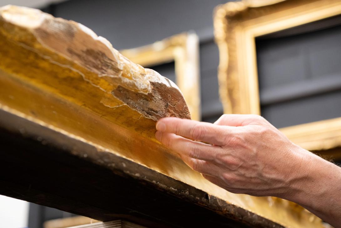 A photograph of an outstretched hand inspecting damaged sections of a gilded frame.