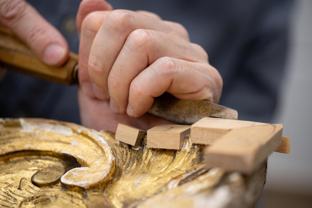This photograph depicts a pair of hands using a chisel to hand-carve details into a gilded frame.