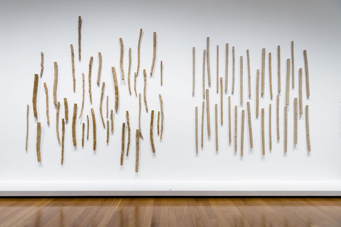 A sculptural work installed on a white gallery wall, taking the form of many gum and bamboo branches made from mud-coloured clay.
