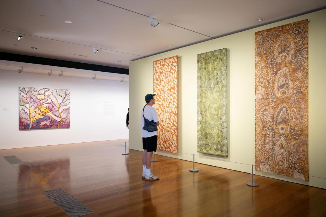 An installation view of textile works by Australian First Nations artists installed on a yellow wall in a gallery space.