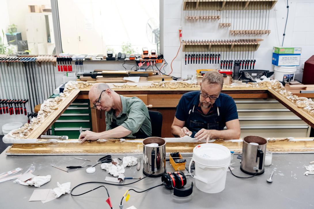 A photograph in which two male conservators sit inside a huge empty frame for an artwork, doing conservation and repair work.
