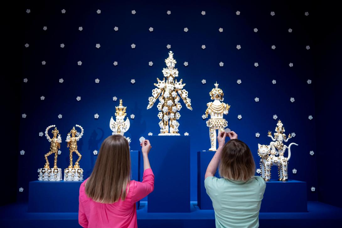 Two gallery visitors dressed in pink and blue each hold up a paper flower in offering to five ceramic deities, white and gold against a dark blue background.