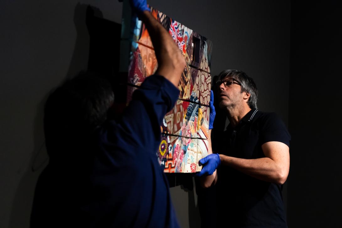 Two masculine-presenting people in a dark gallery space hang a painting on a black wall