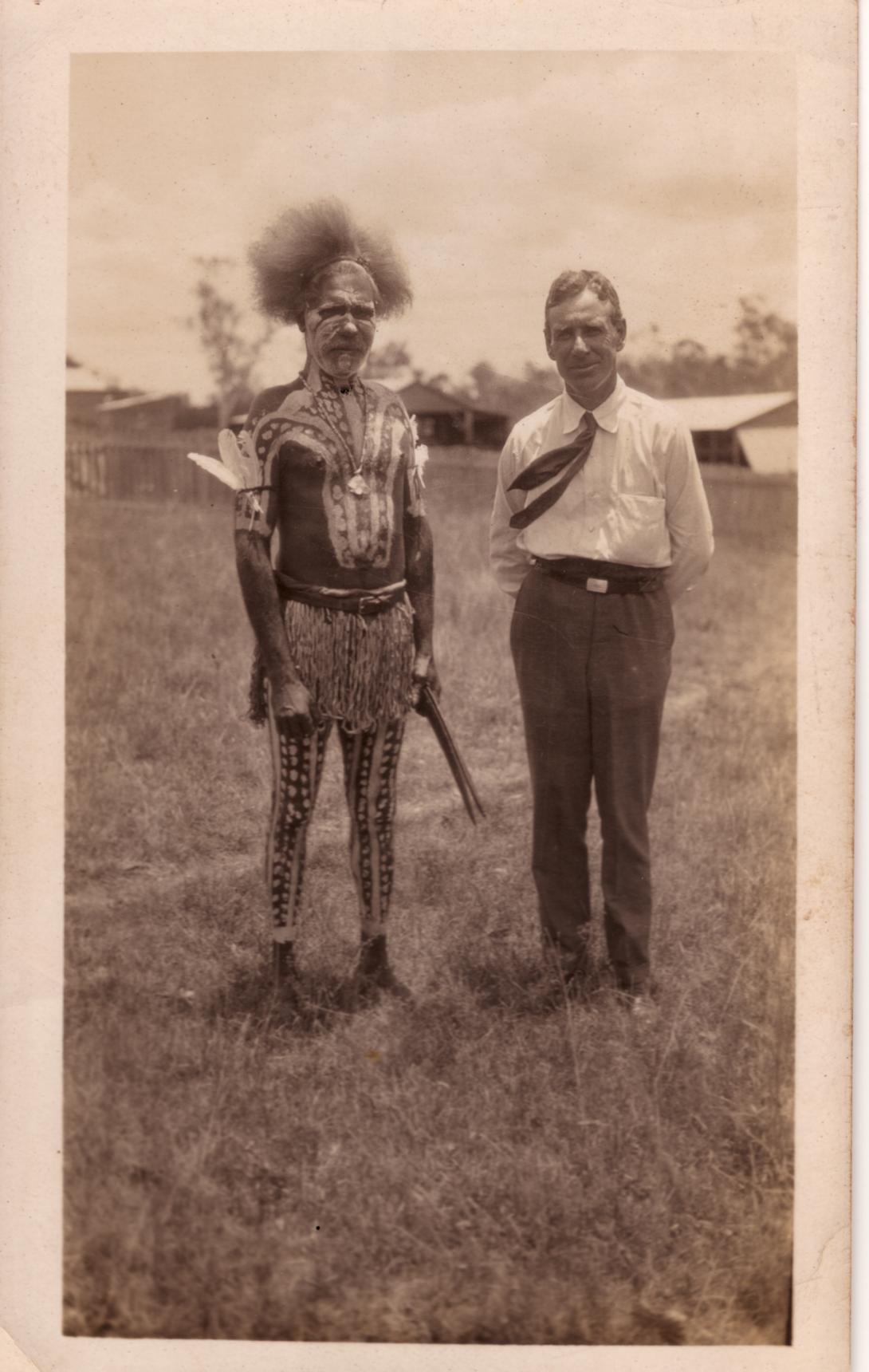 A sepia-toned photo of an Australian Indigenous man in traditional dress standing beside a white man in western dress; they stand outside in an open field