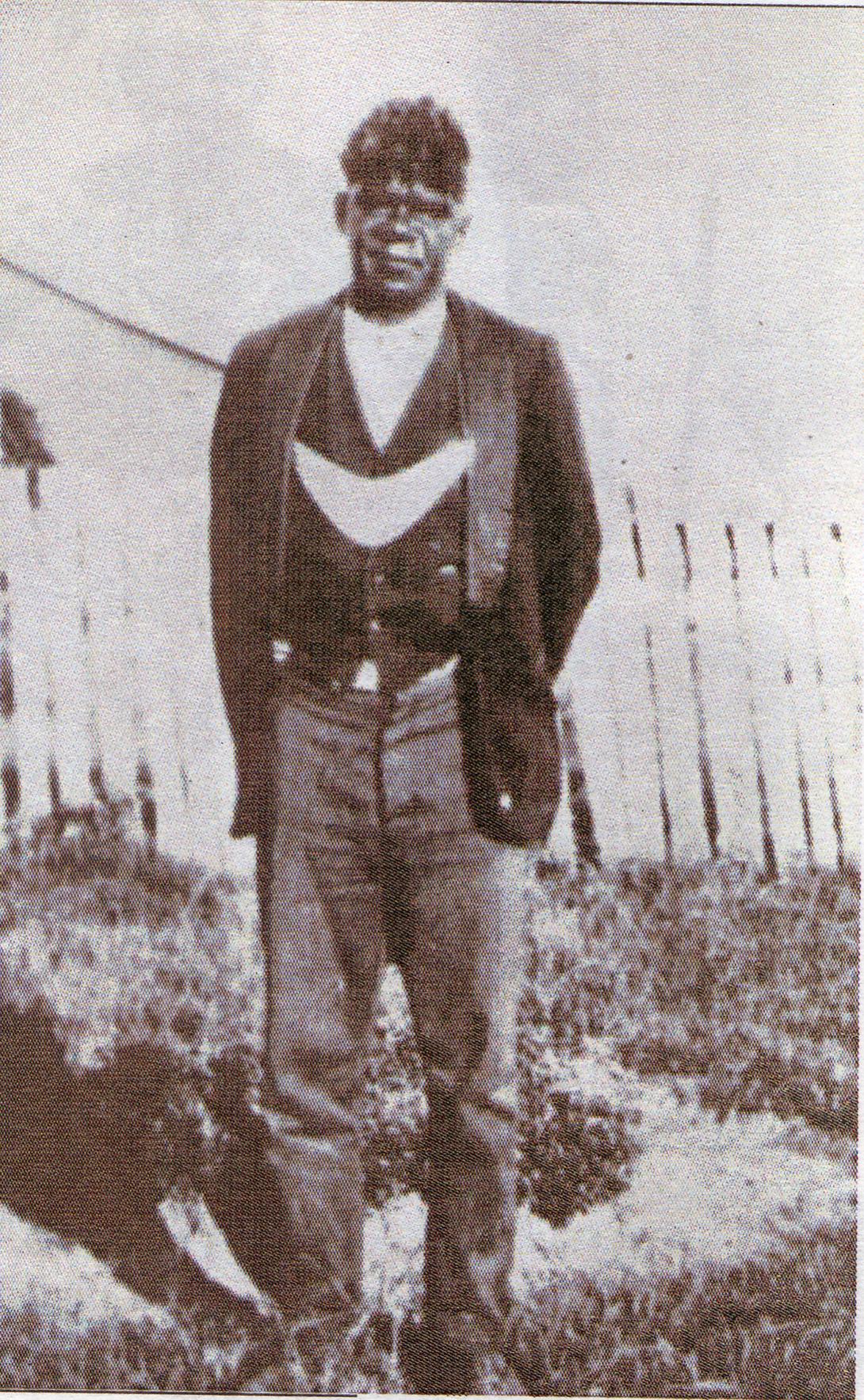 A black-and-white photograph of an Australian Indigenous man wearing a boomerang-shaped 'king plate' around his neck, over his suit