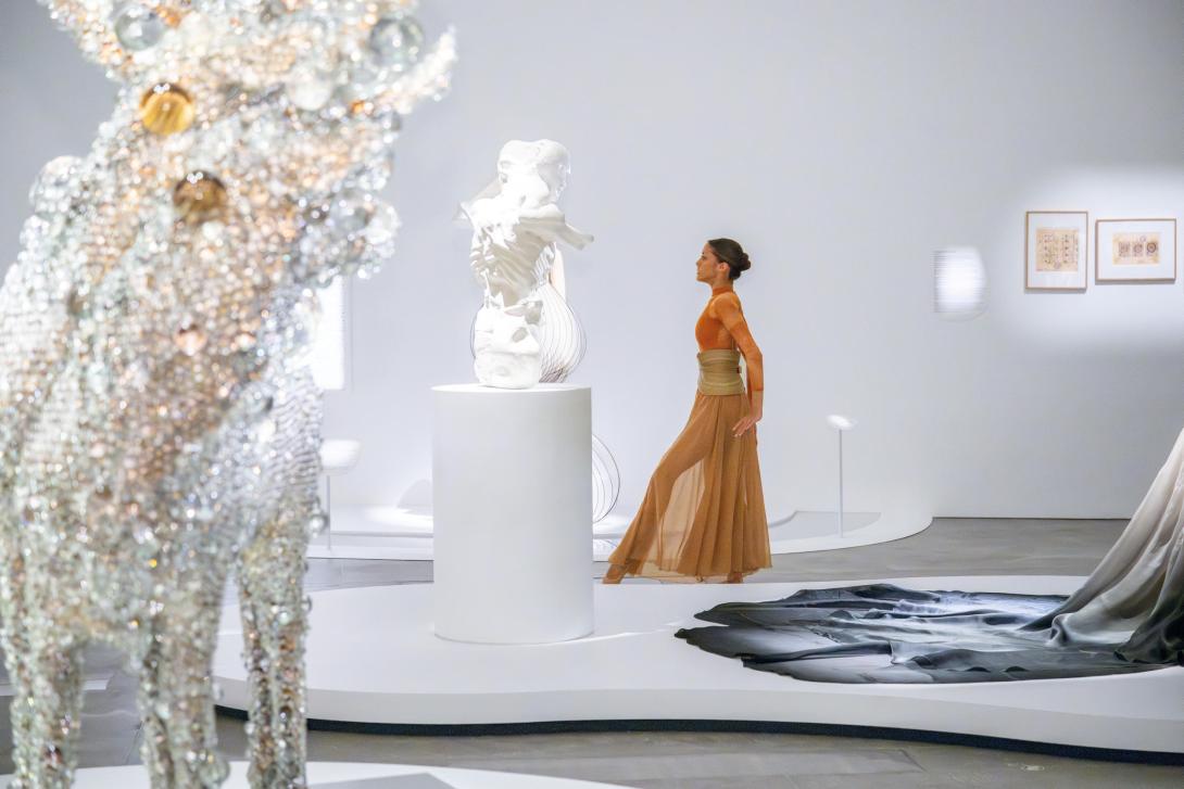 Installation view of a bright gallery space, with a ballet dancer in a floaty, coffee-coloured dress moving between the artworks