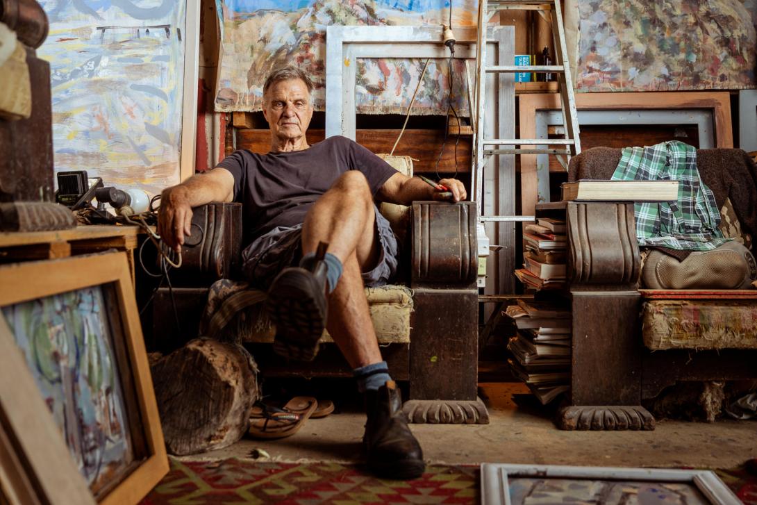 A photograph of a man sitting on a chair in his artist's studio, surrounded by works in progress, supplies and furniture.